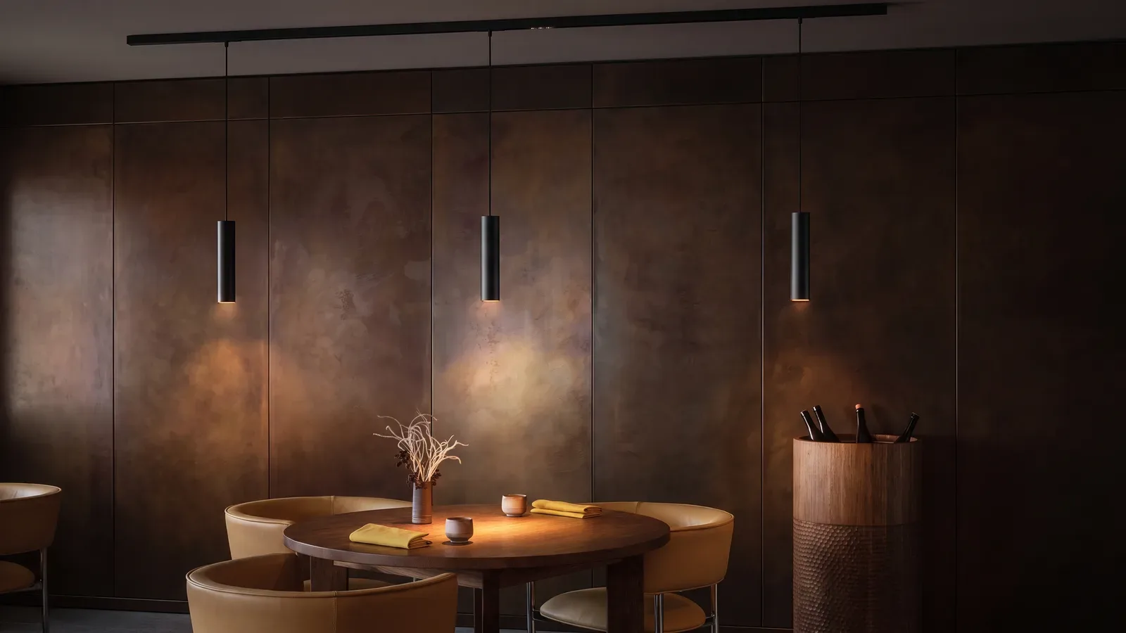 Restaurante de paneles oscuros en tono bronce con un carril magnético negro en el techo y tres colgantes cilíndricos negros: el central cae sobre una mesa redonda con sillones de cuero claro y los otros dos iluminan los paneles.